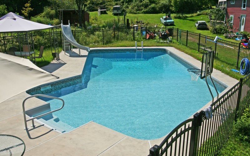 This is a photo of a Lazy L style custom inground swimming pool with a black fence, steps and water slide.