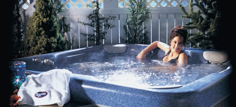 Woman relaxing in a bubbling Saratoga Spa hot tub with steam rising, featuring a mosaic candle holder and towel on the edge